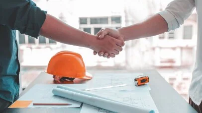 Aperto de mão sobre uma mesa de obra com capacete laranja e plantas de projeto